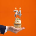 Hand holding birthday cupcake with number 74 candle - background orange