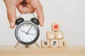 Hand holding black analog alarm clock and pyramid of wooden cubes with KPI icon concept
