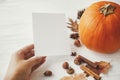 Hand holding blank card on background of pumpkin, autumn leaves, anise, acorns on white wood. Autumn greeting card mock up.