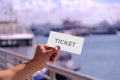 Hand holding blank mockup for ticket or card design, at a pier. Cruise ferry and ships in the background, defocused. Bright sunny