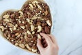 Hand holding Brazil nut and heart-shaped bowl full of mixed nuts isolated on white marble background, top view Royalty Free Stock Photo