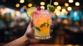 Hand holding a colorful tropical cocktail adorned with lemon and berries, with a backdrop of glowing bokeh lights in a vibrant bar