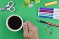 Hand holding a cup of coffee surrounded with stationery tools over green background Royalty Free Stock Photo