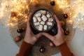 Hand holding cup of hot chocolate with marshmallows decorated Christmas baubles and defocused lights on gray wooden background. Royalty Free Stock Photo