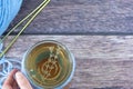 Hand holding a cup of tea and blue knitting crochet yarn with needles on wooden table, top view