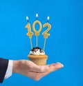 Hand holding a cupcake with the number 102 candle - Birthday on blue background