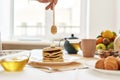 Hand holding dipper, adding honey on top of stack of sweet tasty pancakes, Teapot, croissants and various fruits served