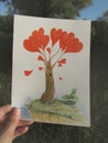 A hand is holding a drawing of a fantastic tree with red hearts in the light of the rising sun