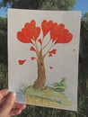 A hand is holding a drawing of a fantastic tree with red hearts, on which a bug sits, in the light of the rising sun