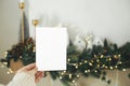 Hand holding empty greeting card on background of stylish christmas houses, fir branches with golden lights and tree decorations Royalty Free Stock Photo