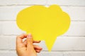 Hand holding an empty yellow hand-cuted paper speech bubble on white textured brick wall background. .