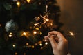 Hand holding firework against christmas tree lights in dark room. Happy New Year! Merry Christmas! Burning sparkler in female hand Royalty Free Stock Photo