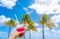 Hand holding a glass with cocktail and straw and Beautiful Palm trees in blue sky background Royalty Free Stock Photo