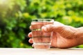 Hand holding glass of fresh drinking water on wooden tabletop on blurred green nature background