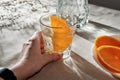 Hand holding glass with orange infused water, bottle, sliced oranges on beige table with aesthetic sunlight shadows, vegan, Royalty Free Stock Photo