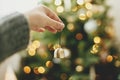 Hand holding golden bell on background of illuminated christmas tree with golden lights. Woman decorating christmas tree and Royalty Free Stock Photo