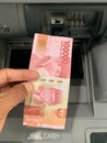 Hand holding a 100,000 IDR banknote in front of an ATM machine Royalty Free Stock Photo