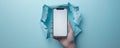 A hand holding an iPhone with a blank white screen emerging through torn blue paper background Royalty Free Stock Photo
