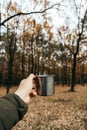 Hand holding an iron cup with a hot drink on a background of autumn forest. vertical photo. mood of adventure and