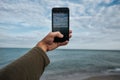 Hand holding mobile phone and take a photo landscape scenery of sea wave with white bubble splashing on beach with blue Royalty Free Stock Photo