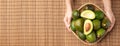 Hand holding organic avocados half cut with seed and whole fruit in basket on bamboo mat on table background.Healthy super foods