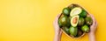 Hand holding organic avocados half cut with seed and whole fruit in basket on yellow table background.Healthy super foods for diet