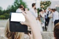 Hand holding phone with empty screen and taking photo of gorgeous bride and stylish groom with guests at wedding reception in Royalty Free Stock Photo