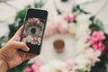 Hand holding phone and taking photo of coffee drink in beautiful pink and white peonies frame on table, flat lay. Good morning Royalty Free Stock Photo