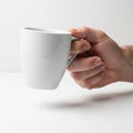A hand is holding a plain white ceramic mug with the handle towards the viewer against a clean, Royalty Free Stock Photo