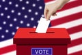 Hand holding and putting voting paper in ballot voting box with USA flag in background. Royalty Free Stock Photo