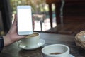 A hand holding and showing a white mobile phone with blank desktop screen with coffee cups on wooden table in cafe Royalty Free Stock Photo