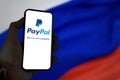 Hand holding a mobile phone with the Paypal app on the screen with the Russian flag blurred in the background