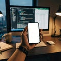 A hand holding a smartphone with a blank screen in a modern workspace featuring a computer, keyboards, and warm lighting Royalty Free Stock Photo