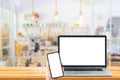 Hand holding smartphone and Used laptop with blank white screen on wooden table and blurry image of coffee cafe in background.