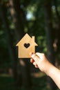 Hand holding a sunlit wooden house model against a forest background.