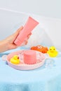 A hand holding a tube of pink cosmetics, a jar of lotion, silicone bath toys, a bath sponge, ceramic tray and ice are placed on a Royalty Free Stock Photo