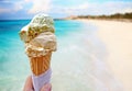 Hand is holding the vanilla ice cream. In the background is blue sky, sea nd sandy beach. This is situated in tropical resort