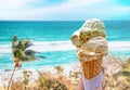 Hand is holding the vanilla ice cream. In the background is blue sky, sea nd sandy beach. There are palms