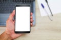 Hand holding white blank screen smartphone above blurred office wooden desk with laptop computer,paper and pens Royalty Free Stock Photo