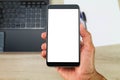 Hand holding white blank screen smartphone above blurred office wooden desk with laptop computer,paper and pens Royalty Free Stock Photo