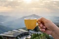 Hand holding a white cup of hot espresso coffee mugs and nature view of the mountain landscape in the morning with sunlight