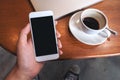 Hand holding white mobile phone with blank black desktop screen with coffee cup and laptop on wooden table in cafe Royalty Free Stock Photo