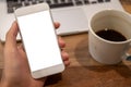 Hand holding white mobile phone with blank desktop screen with coffee cup and laptop on wooden table in cafe Royalty Free Stock Photo