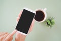 Hand holding white smartphone with clipping path on touchscreen, top view, and blurred fake small tree and cup of coffee