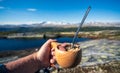 Hand holding a wooden cup of spaghetti and salty sausage outdoors towards mountain scenery.
