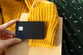 Hand holds black plastic credit card blank. Yellow wool knitting sweater, green skirt and craft paper bag. Shopping online