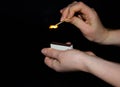 A hand holds a burning match on a black background. A wooden match burns in the hands of a macro. Igniting a match on a box. Smoke