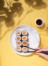 A hand holds chopsticks with Japanese sushi rolls on a white plate and soy sauce on a yellow background with a hard shadow brunch Royalty Free Stock Photo