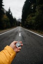 A hand holds a compass on a mountain road surrounded by forest