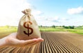 A hand holds out a dollar money bag on a background of a farm field. Lending farmers and agricultural enterprises for purchase